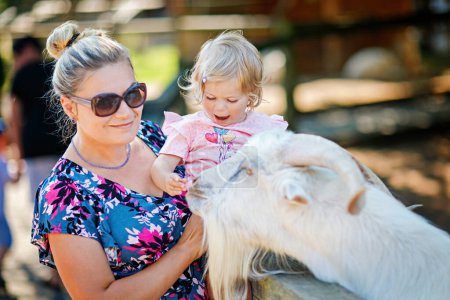 Photo for Adorable cute toddler girl and young mother feeding little goats and sheeps on a kids farm. Beautiful baby child petting animals in petting zoo. Woman and daughter together on family weekend vacations - Royalty Free Image