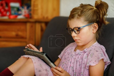 Foto de Niña positiva, sosteniendo la tableta en sus manos. Niño preescolar con anteojos jugando juegos y aprendiendo con diferentes aplicaciones - Imagen libre de derechos
