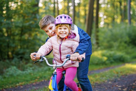 Photo for Cute little preschool girl in safety helmet riding bicycle. School kid boy, brother teaching happy healthy sister child cycling and having fun with learning bike. Active siblings family outdoors - Royalty Free Image