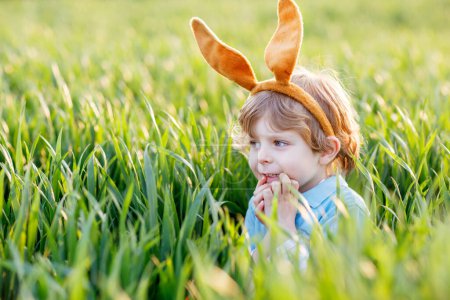 Foto de Niño lindo con orejas de conejo divirtiéndose con la tradicional caza de huevos de Pascua en el cálido día soleado, al aire libre. Celebrando las vacaciones de Pascua. Encontrar niños pequeños, huevos coloridos en hierba verde. - Imagen libre de derechos