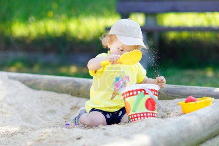 Téléchargez les photos : Petite fille mignonne jouant dans le sable sur une aire de jeux extérieure. Beau bébé qui s'amuse par une chaude journée ensoleillée d'été. Heureux enfant en bonne santé avec des jouets de sable et des vêtements de mode colorés - en image libre de droit