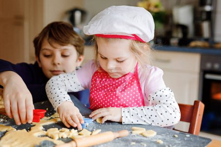 Photo for Cute little toddler girl and preteen kid boy baking Easter cookies at home indoors. Children, siblings with apron and chef hat cut out bunny cookie in domestic kitchen. Brother and sister in love. - Royalty Free Image
