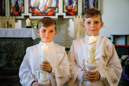 Téléchargez les photos : Deux petits garçons recevant sa première sainte communion. Des enfants heureux tenant une bougie du baptême. Tradition en curch catholique. Des enfants dans une église près de l'autel. Frères et sœurs, frères et sœurs en robes blanches - en image libre de droit