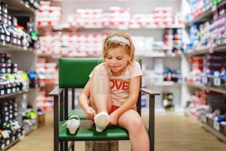 Téléchargez les photos : Happy Little Girl sélectionne et achète soigneusement de nouvelles chaussures dans un magasin, en essayant diverses options pour trouver la paire parfaite. Enfant préscolaire excité - en image libre de droit