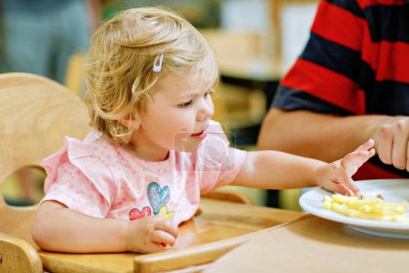 Photo for Toddler girl eating healthy vegetables and unhealthy french fries potatoes. Cute happy baby child taking food from parents dish in restaurant. Father eating in fast food restaurant with daughter. - Royalty Free Image