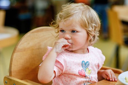 Foto de Adorable niña comiendo verduras saludables y papas fritas poco saludables. Lindo bebé feliz niño tomando comida del plato en la guardería o la cantina del vivero - Imagen libre de derechos