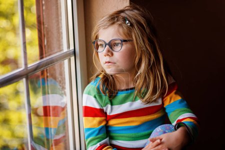 Foto de Adorable niña preescolar con anteojos sentados junto a la ventana. Niño reflexivo mirando hacia fuera. Chico solitario - Imagen libre de derechos