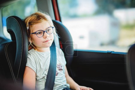 Photo for Little Preschool Girl Sitting in Her Car Seat. Happy Child with Eyeglasses looking out of the window. Smiling kid on the Way to Family Vacations during Traffic Jam - Royalty Free Image