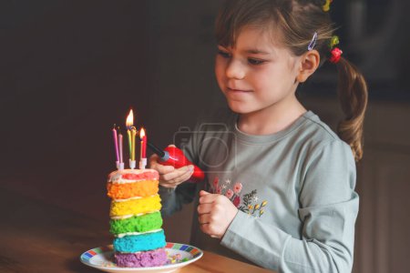 Foto de Happy little preschool girl celebrating birthday. Cute smiling child with homemade rainbow cake, indoor. Happy healthy toddler blowing six candles on cake - Imagen libre de derechos