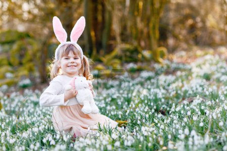 Téléchargez les photos : Adorable petite fille avec des oreilles de lapin de Pâques tenant peluche douce dans la forêt de printemps le jour ensoleillé, à l'extérieur. Mignon enfant heureux avec beaucoup de fleurs de chute de neige. Printemps, concept de vacances chrétiennes - en image libre de droit
