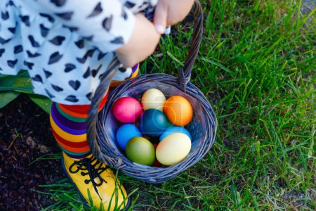 Photo for Close-up of legs of toddler girl with colorful stockings and shoes and basket with colored eggs. Child having fun with traditional Easter eggs hunt, outdoors. Celebration of christian holiday - Royalty Free Image