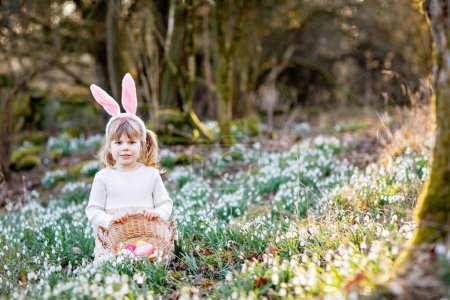 Photo for Little girl making Easter egg hunt in spring forest on sunny day, outdoors. Cute happy child with lots of snowdrop flowers and colored eggs. Springtime, christian holiday concept - Royalty Free Image