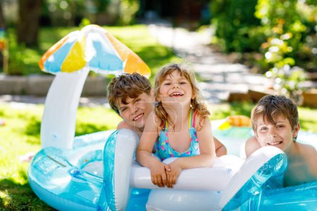 Téléchargez les photos : Trois enfants, deux garçons et une petite fille éclaboussent dans une piscine extérieure en été. Enfants heureux, frères et sœurs jouant dans la piscine gonflable de gomme appréciant le temps ensoleillé dans le jardin domestique - en image libre de droit