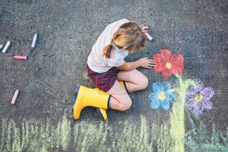 Téléchargez les photos : Jolie petite fille et fleurs peintes avec des craies colorées sur asphalte. Joyeux enfant d'âge préscolaire s'amuser avec la peinture tableau à la craie. Loisirs créatifs pour les enfants, dessin et peinture - en image libre de droit