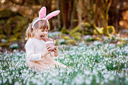 Foto de Niña feliz con orejas de conejo de Pascua comiendo figura de chocolate en el bosque de primavera en el día soleado, al aire libre. Lindo niño con un montón de flores de nieve. Primavera, concepto de vacaciones cristianas - Imagen libre de derechos