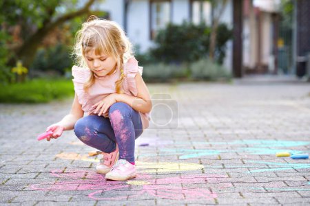 Téléchargez les photos : Petite peinture de fille préscolaire avec des craies colorées fleurs sur le sol sur la cour arrière. Bébé heureux positif dessin d'enfant et la création d'images sur asphalte. Activité créative pour enfants en plein air en été - en image libre de droit