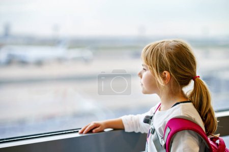 Foto de Niña en el aeropuerto esperando el embarque en la ventana grande. Cute Kid se para en la ventana contra el telón de fondo de los aviones. Mirando hacia adelante a la partida para unas vacaciones de verano familiares. - Imagen libre de derechos