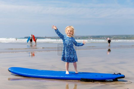 Téléchargez les photos : Little cute toddler girl at the Ballybunion surfer beach, having fun on surfboard for the first time, west coast of Ireland. Happy child enjoying Irish summer with family - en image libre de droit
