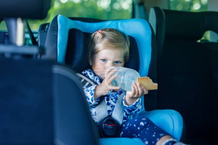 Photo for Adorable baby girl with blue eyes sitting in car safety seat. Toddler child going on family vacations and jorney. Smiling happy child during traffic jam, drinking milk from bottle and eating bisquit. - Royalty Free Image