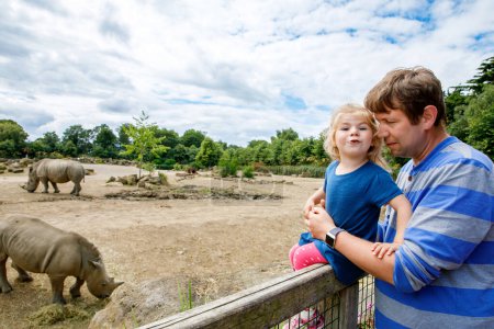 Photo for Cute adorable toddler girl and father watching wild rhinos in zoo. Happy baby child, daughter and dad, family having fun together with animals safari park on warm summer day. Ireland. - Royalty Free Image