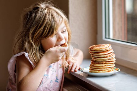Foto de Pequeña niña preescolar feliz con una gran pila de panqueques para el desayuno. Niño positivo comiendo comida casera saludable por la mañana - Imagen libre de derechos
