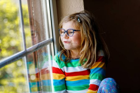 Téléchargez les photos : Adorable petite fille d'âge préscolaire avec des lunettes assis près de la fenêtre. Enfant réfléchi regardant dehors. Enfant solitaire - en image libre de droit