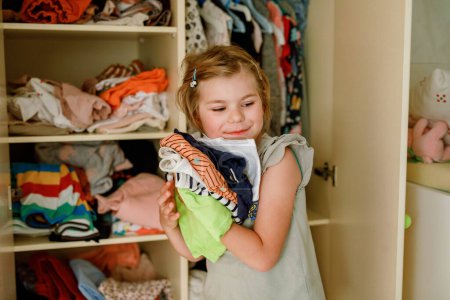Téléchargez les photos : Petite écolière debout près d'une armoire avec des vêtements. Enfant d'âge préscolaire mettre des chemises et de prendre la décision pour chemise à porter. Les enfants s'habillent le matin pour l'école - en image libre de droit