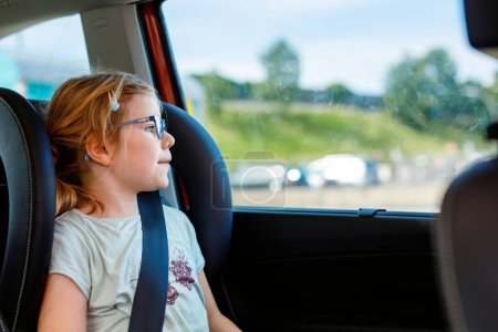 Photo for Little Preschool Girl Sitting in Her Car Seat. Happy Child with Eyeglasses looking out of the window. Smiling kid on the Way to Family Vacations during Traffic Jam - Royalty Free Image