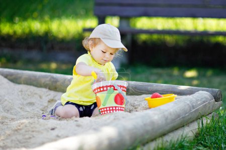 Foto de Linda niña jugando en la arena en el patio al aire libre. Hermoso bebé que se divierte en el soleado cálido día soleado de verano. Feliz niño sano con juguetes de arena y en ropa de moda colorida - Imagen libre de derechos