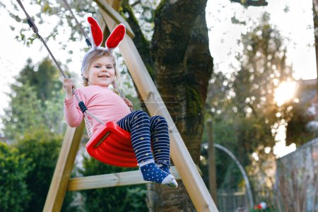 Photo for Happy beautiful little toddler girl with red Easter bunny ears having fun on swing in domestic garden. Cute healthy child swinging under blooming trees on sunny spring day. Baby laughing and crying. - Royalty Free Image
