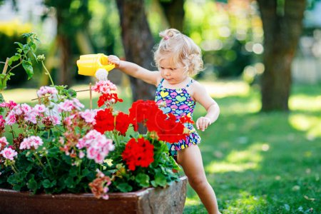 Photo for Cute little baby girl in colorful swimsuit watering plants and blossoming flowers in domestic garden on hot summer day. Adorable toddler child having fun with playing with water and can. - Royalty Free Image