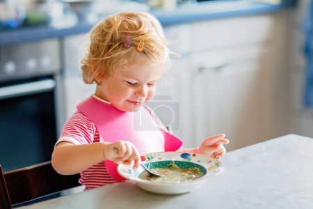 Téléchargez les photos : Adorable petite fille mangeant de la soupe de nouilles aux légumes cuillère. Alimentation saine, enfant, concept d'alimentation et de développement. Enfant tout-petit mignon avec cuillère assis dans une chaise haute et apprenant à manger par lui-même. - en image libre de droit