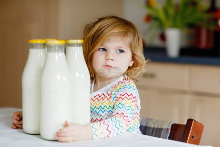 Photo for Adorable toddler girl drinking cow milk for breakfast. Cute baby daughter with lots of bottles. Healthy child having milk as health calcium source. Kid at home or nursery in the morning - Royalty Free Image