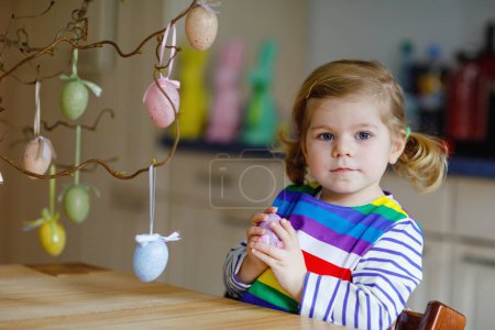 Téléchargez les photos : Jolie petite fille tout-petit décorant la branche d'arbre avec des œufs en plastique pastel colorés. Bébé enfant heureux s'amuser avec des décorations de Pâques. Adorable enfant souriant en bonne santé en profitant des vacances en famille. - en image libre de droit