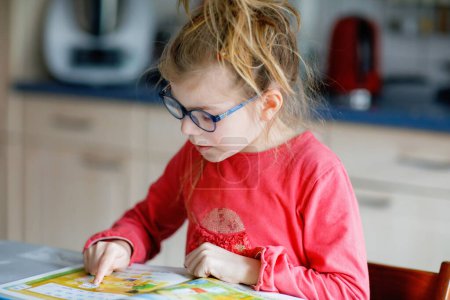 Téléchargez les photos : Petite fille avec des lunettes apprenant à lire. Enfant de l'école primaire lit un livre à la maison, faire des devoirs scolaires - en image libre de droit