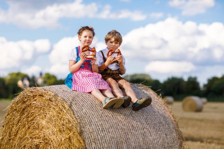 Photo for Two kids in traditional Bavarian costumes in wheat field. German children eating bread and pretzel during Oktoberfest. Boy and girl play at hay bales during summer harvest time in Germany - Royalty Free Image