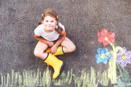 Téléchargez les photos : Jolie petite fille et fleurs peintes avec des craies colorées sur asphalte. Joyeux enfant d'âge préscolaire s'amuser avec la peinture tableau à la craie. Loisirs créatifs pour les enfants, dessin et peinture - en image libre de droit