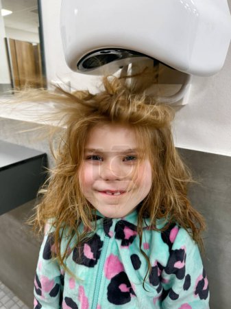 Foto de Pequeña chica sonriente seca los pelos con secador de pelo después de la piscina vistining. Niño feliz con cabellos largos y rubios - Imagen libre de derechos