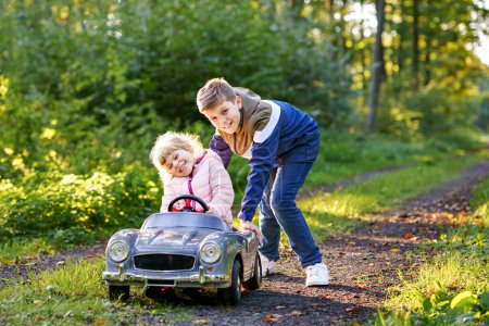 Téléchargez les photos : Deux enfants heureux jouant avec une grande vieille voiture jouet dans la forêt d'automne, à l'extérieur. Petit garçon poussant et conduisant voiture avec petite fille tout-petit, jolie soeur à l'intérieur. Des enfants riants et souriants. Belle famille. - en image libre de droit