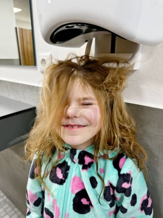 Téléchargez les photos : Petite fille souriante sèche les cheveux avec sèche-cheveux après avoir vissé la piscine. Enfant heureux avec de longs cheveux blonds - en image libre de droit