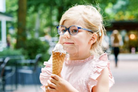 Foto de Little preschool girl with glasses eating ice cream in waffle cone on sunny summer day. Happy toddler child eat icecream dessert. Sweet food on hot warm summertime days. - Imagen libre de derechos
