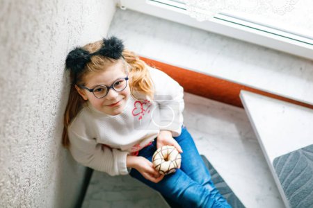 Téléchargez les photos : Bonne petite écolière mangeant un beignet sucré à la maison. Enfant blond avec beignet. Enfant mignon avec des lunettes et des beignets alimentaires sucrés malsains - en image libre de droit