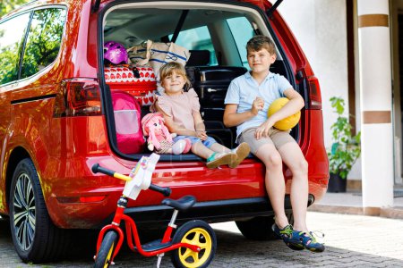Photo for Two children, school boy and preschool girl sitting in car trunk before leaving for summer vacation with parents. Happy kids, siblings, brother and sister with suitcases and toys going on journey. - Royalty Free Image