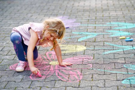 Photo for Little preschool girl painting with colorful chalks flowers on ground on backyard. Positive happy toddler child drawing and creating pictures on asphalt. Creative outdoors children activity in summer - Royalty Free Image