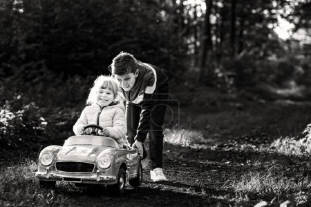 Téléchargez les photos : Deux enfants heureux jouant avec une grande vieille voiture jouet dans la forêt d'automne, à l'extérieur. Petit garçon poussant et conduisant voiture avec petite fille tout-petit, jolie soeur à l'intérieur. Des enfants riants et souriants. Belle famille. - en image libre de droit