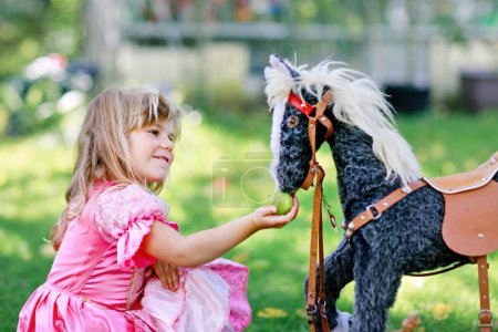Téléchargez les photos : Petite fille préscolaire étreignant avec jouet de cheval à bascule. Joyeux enfant en robe de princesse le jour ensoleillé d'été dans le jardin. Fille en amour avec son favori vieux jouet vintage animal - en image libre de droit