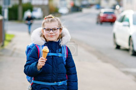 Foto de Linda niñita camino a casa desde la escuela. Niño feliz saludable con anteojos con cono de helado. Niño sonriente con bolsa de escuela y helado en la calle de la ciudad, al aire libre - Imagen libre de derechos