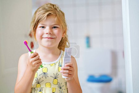 Téléchargez les photos : Cute little girl with a toothbrush and toothpaste in her hands cleans her teeth and smiles. Happy preschool child brushing first teeth - en image libre de droit