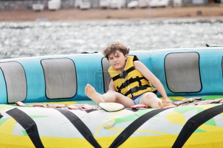 Foto de Niño joven exuda alegría mientras monta un tubo inflable remolcado por un barco en el océano. Niño de la escuela feliz divirtiéndose en el parque acuático de aventura en el mar - Imagen libre de derechos