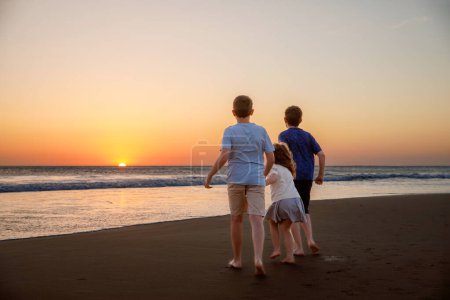 Photo for Three kids silhouettes running and jumping on beach at sunset. happy family, two school boys and one little preschool girl. Siblings having fun together. Bonding and family vacation - Royalty Free Image